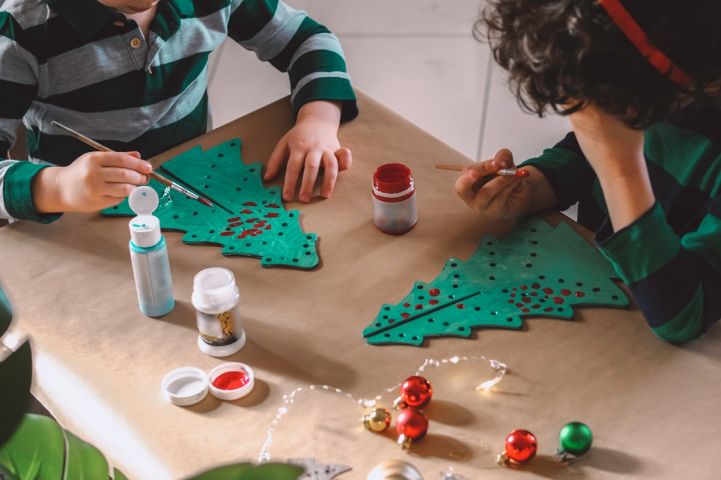 Dzieci malują farbami zielone ozdoby w kształcie choinek przy stole; obok stoją pojemniki z farbą i leżą czerwone bombki.