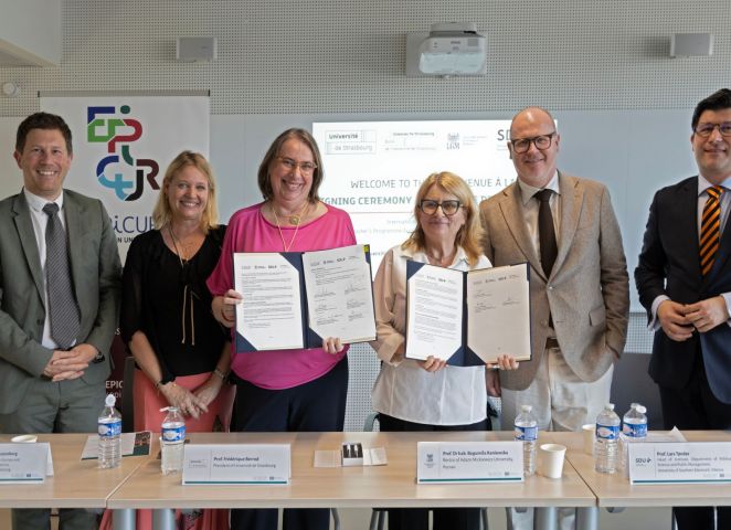 Grupa osób stoi za stołem i prezentuje podpisane dokumenty podczas wydarzenia EPICUR; w tle widoczne są banery oraz ekran z informacją o ceremonii podpisania.
