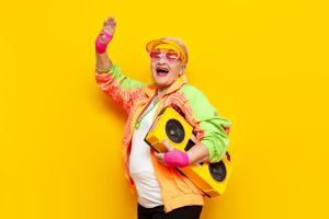 Uśmiechnięta starsza kobieta w jaskrawym, kolorowym stroju i różowych okularach unosi rękę i trzyma żółty boombox na intensywnie żółtym tle.