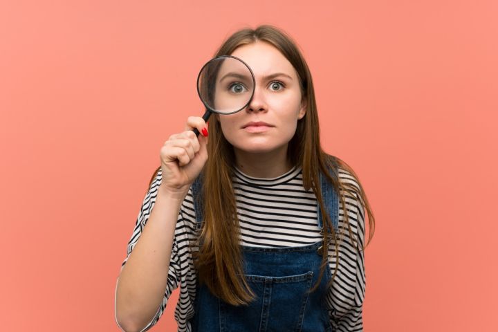 Młoda kobieta patrzy przez lupę na różowym tle.
