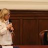 Osoba stoi przy stole i mówi do mikrofonu, zwracając się do siedzącej obok osoby podczas panelu w eleganckim wnętrzu.