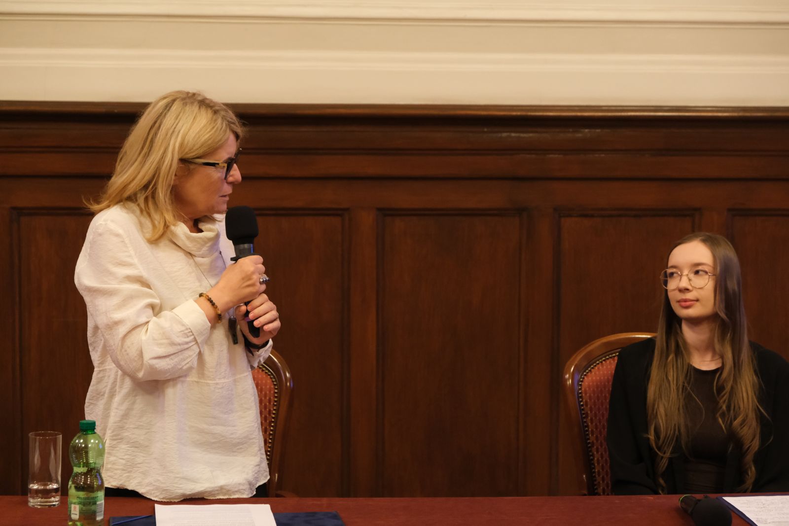 Osoba stoi przy stole i mówi do mikrofonu, zwracając się do siedzącej obok osoby podczas panelu w eleganckim wnętrzu.