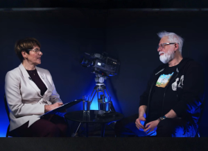 Rozmowa studyjna: Małgorzata Rybczyńska prowadząca wywiad i Tomasz Raczek siedzą naprzeciw siebie przy stoliku, między nimi kamera na statywie; ujęcie w ciemnym studiu z niebieskim oświetleniem.