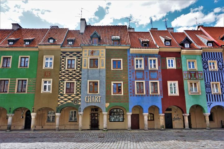 Kolorowe kamienice z ozdobnymi fasadami i podcieniami na Starym Rynku w Poznaniu; pastelowe elewacje z geometrycznymi i historycznymi wzorami, czerwone dachówki i brukowana ulica na pierwszym planie.