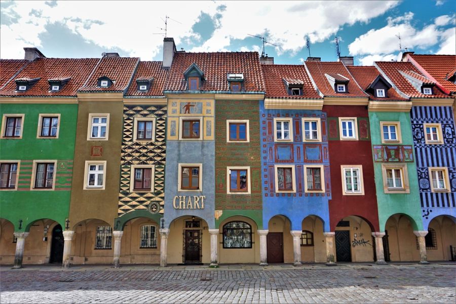 Kolorowe kamienice z ozdobnymi fasadami i podcieniami na Starym Rynku w Poznaniu; pastelowe elewacje z geometrycznymi i historycznymi wzorami, czerwone dachówki i brukowana ulica na pierwszym planie.