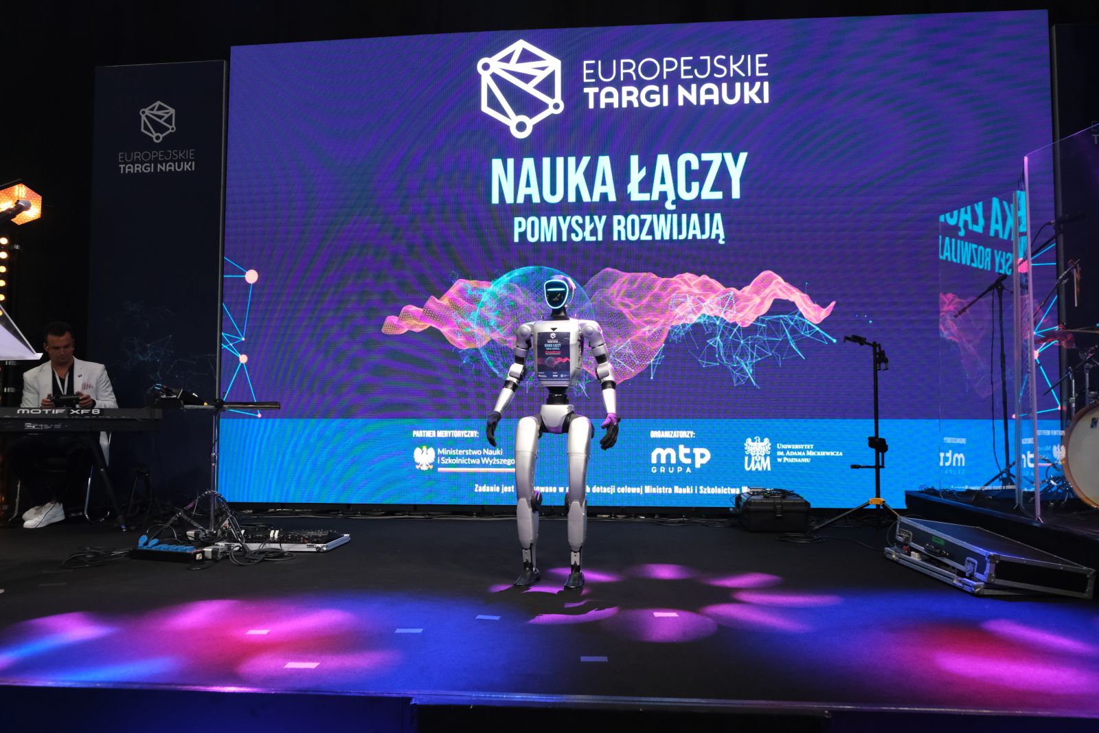 Humanoidalny robot stoi na scenie przed dużym ekranem z napisem „Europejskie Targi Nauki – Nauka łączy, pomysły rozwijają”, wokół widoczne są instrumenty muzyczne i oświetlenie sceniczne.