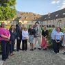 Grupa osób stoi w ogrodzie przed historycznym budynkiem z arkadami i wspólnie pozuje do zdjęcia; w tle widoczna jest rzeźba oraz zieleń.