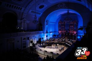 Koncert w Auli UAM. Na scenie chór. W prawym dolnym rogu logo aukcji 33 Finału WOŚP