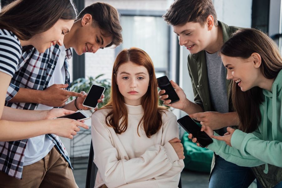 Grupa nastolatków trzymająca smartfony w ręku, wskazująca palcem i naśmiewająca się z dziewczyny siedzącej w ich otoczeniu