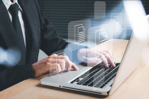 Mężczyzna w garniuturze obsługujący laptopa. Nad klawiaturą unoszące się ikony symbolizujące pliki tekstowe.