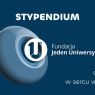 Grafika na granatowym tle promująca stypendium Fundacji „Jeden Uniwersytet”. Po lewej trójwymiarowy pierścień z logo fundacji. Tekst: „STYPENDIUM”, „Fundacja Jeden Uniwersytet”, „od 10 lat w sercu wydarzeń”.