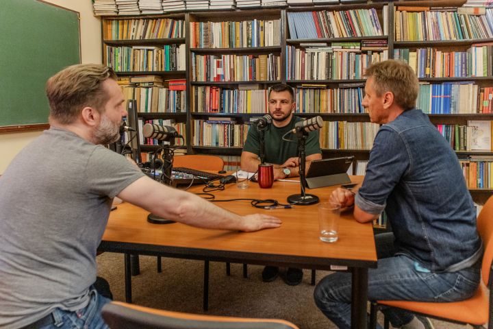 Trzech mężczyzn siedzi przy stole w pomieszczeniu pełnym książek, nagrywając rozmowę przy mikrofonach. Na stole leżą laptopy, mikser dźwięku i szklanki z wodą. W tle widać regały wypełnione książkami oraz tablicę szkolną.