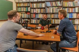Trzech mężczyzn siedzi przy stole w pomieszczeniu pełnym książek, nagrywając rozmowę przy mikrofonach. Na stole leżą laptopy, mikser dźwięku i szklanki z wodą. W tle widać regały wypełnione książkami oraz tablicę szkolną.