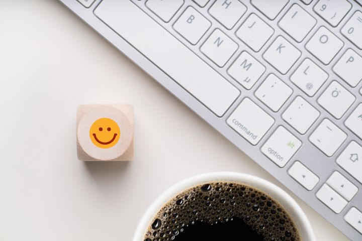 Drewniana kostka z narysowaną żółtą buźką leży na biurku obok klawiatury komputera i kubka czarnej kawy.