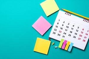 Widok z góry na biurko z kalendarzem na spirali, kolorowymi karteczkami samoprzylepnymi, ołówkiem i pinezkami, na turkusowym tle.