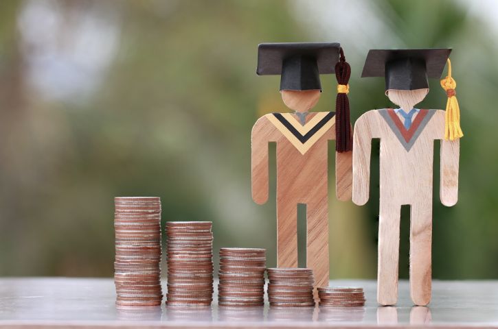 Two wooden figurines of graduates wearing academic caps stand next to several piles of coins of varying heights.