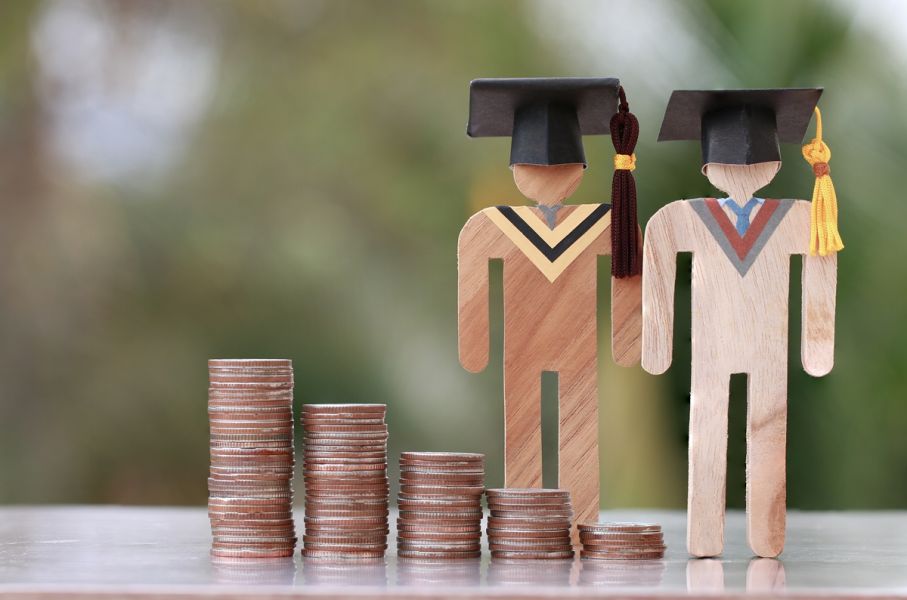 Two wooden figurines of graduates wearing academic caps stand next to several piles of coins of varying heights.