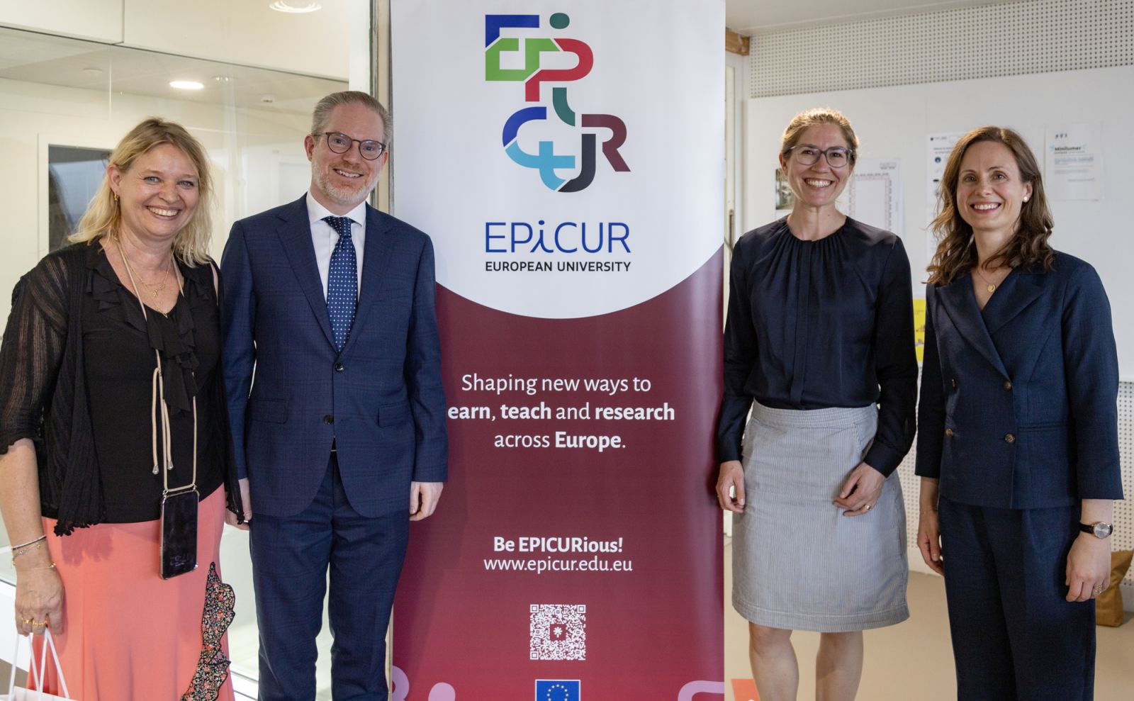 Cztery osoby stoją po obu stronach roll‑upu EPICUR z napisem „European University – Shaping new ways to learn, teach and research across Europe” i pozują do zdjęcia w przestrzeni biurowej.