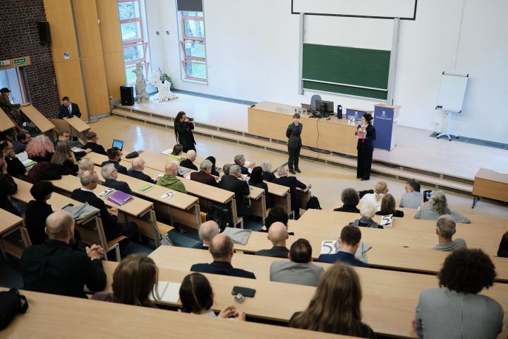 Uczestnicy siedzą w auli wykładowej i słuchają wystąpienia; na dole sali stoją dwie osoby, jedna przemawia do mikrofonu, obok widoczny jest ekran i tablica.