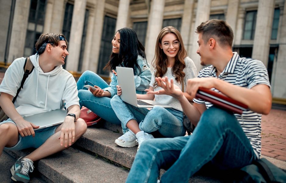 Grupa uśmiechniętych studentów siedząca na schodach. Jedna z osób trzyma laptopa.