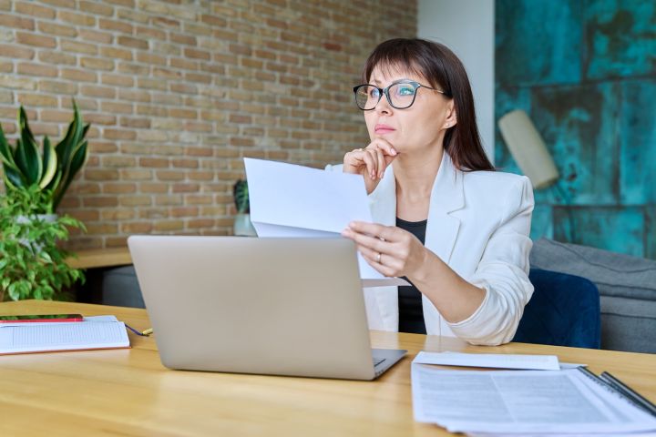 Kobieta w okularach i jasnej marynarce siedzi przy biurku z laptopem, trzyma kartkę papieru i zamyślona patrzy w dal; na stole leżą dokumenty i notatnik.