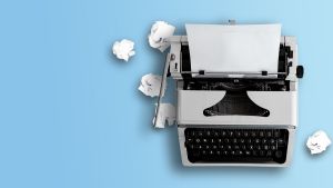 Maszyna do pisania z włożoną kartką papieru na jasnoniebieskim tle; obok leżą zgniecione kartki.