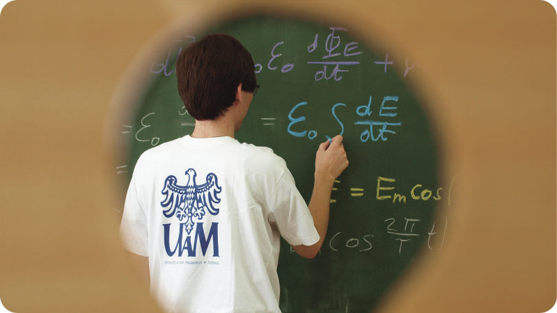 Student z koszulką z logo UAM rozwiązuje zadanie matematyczne, pisząc kredą na tablicy