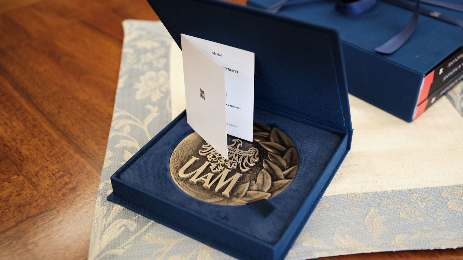 Medal z napisem „UAM” umieszczony w otwartym etui, z dołączoną kartą informacyjną, leży na drewnianym stole.