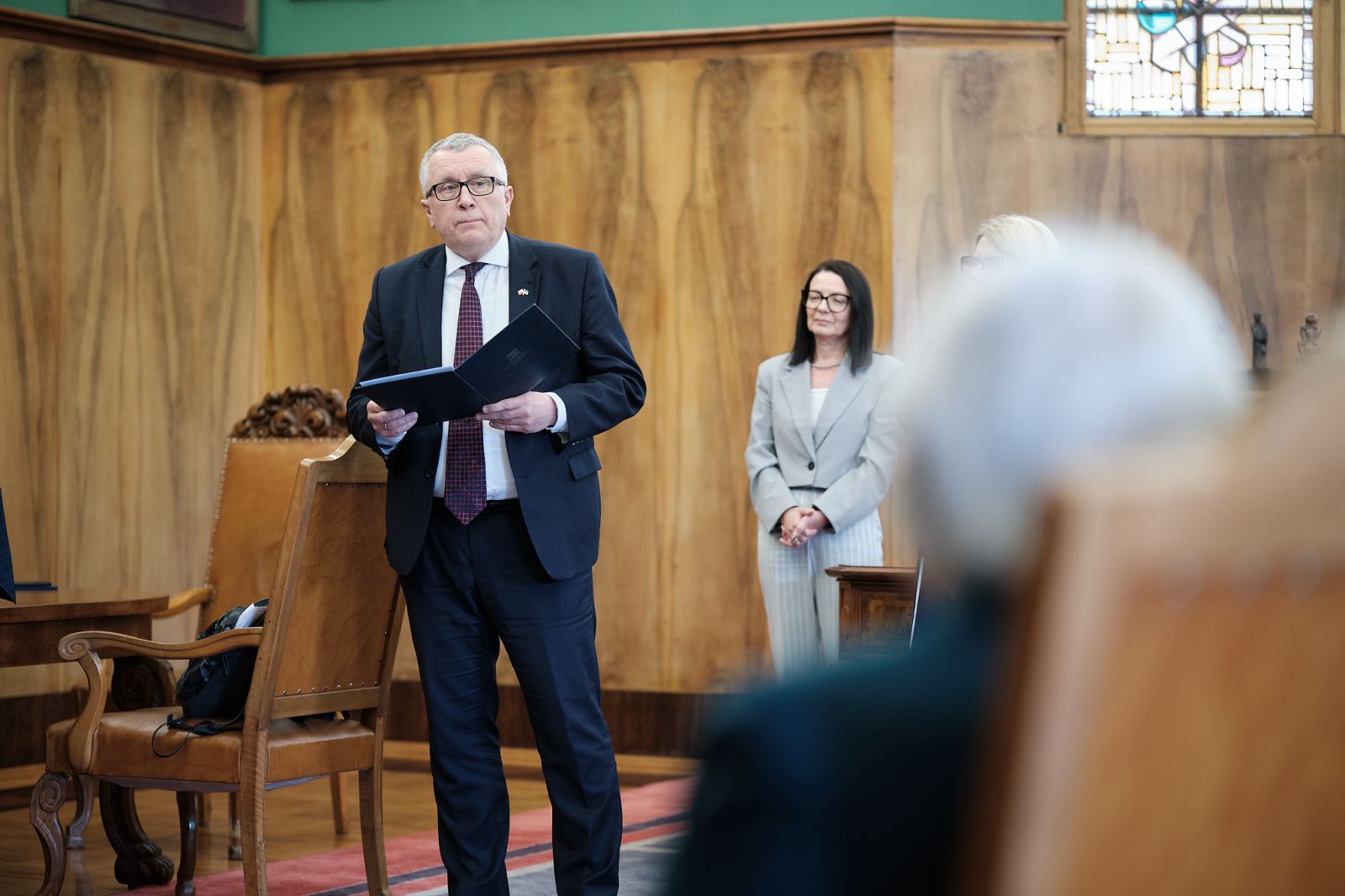 Osoba w garniturze trzyma otwartą teczkę i przemawia do zgromadzonych w eleganckim wnętrzu; w tle stoją uczestnicy spotkania.