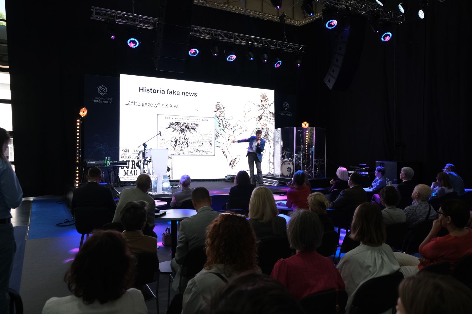 Prelegent stoi na scenie i prowadzi prezentację na temat historii fake news, na dużym ekranie w tle widoczny jest slajd z napisem „Historia fake news” oraz ilustracjami; na widowni siedzą uczestnicy wydarzenia.
