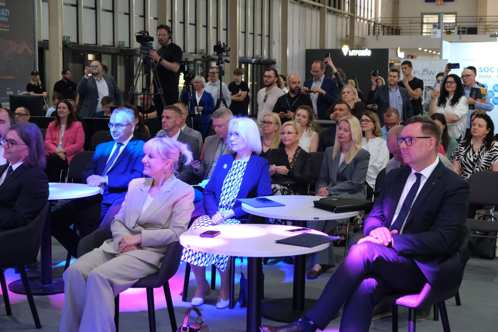 Publiczność siedzi przy stolikach i obserwuje prezentację podczas targów nauki; część osób stoi z tyłu i nagrywa wydarzenie, a w tle widoczne są stoiska i oznaczenia hali wystawowej.