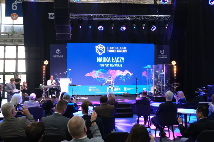 Scena wydarzenia „Europejskie Targi Nauki” z humanoidalnym robotem na środku oraz publicznością siedzącą przy stolikach; na dużym ekranie widoczne jest hasło „Nauka łączy, pomysły rozwijają”.