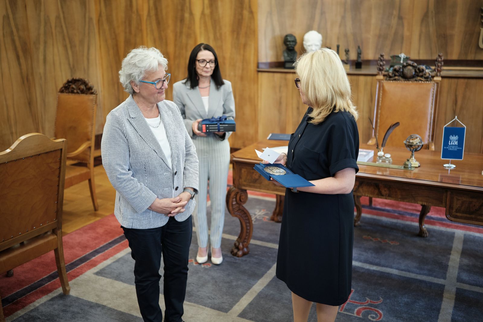 Dwie osoby stoją naprzeciw siebie w reprezentacyjnej sali; jedna trzyma otwarte etui z medalem i kartą informacyjną, w tle widoczna osoba z pudełkiem.