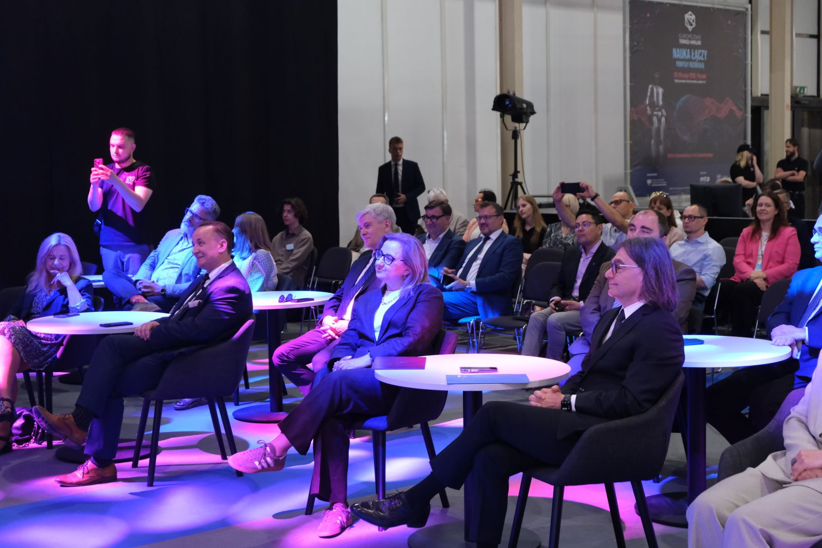 Uczestnicy wydarzenia siedzą przy okrągłych stolikach i słuchają wystąpienia na scenie; część osób nagrywa lub fotografuje wydarzenie, a przestrzeń oświetlona jest kolorowym światłem scenicznym.
