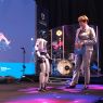 Osoba stoi na scenie obok humanoidalnego robota i trzyma dokument, w tle widoczny jest zestaw perkusyjny oraz ekran z grafiką Europejskich Targów Nauki.
