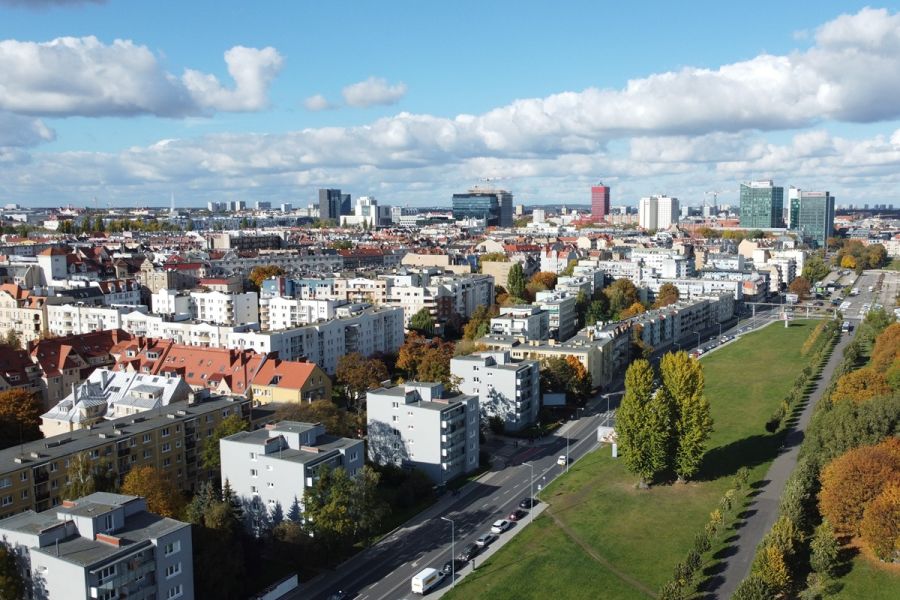 Panoramiczny widok na zabudowę miejską Poznania z lotu ptaka: osiedla mieszkaniowe, szeroka ulica i pas zieleni, w tle centrum z nowoczesnymi biurowcami.