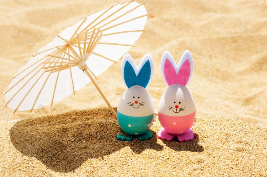 Dwie kolorowe figurki wielkanocnych zajączków stojące na piasku, obok mały papierowy parasol plażowy, w słonecznej scenerii.