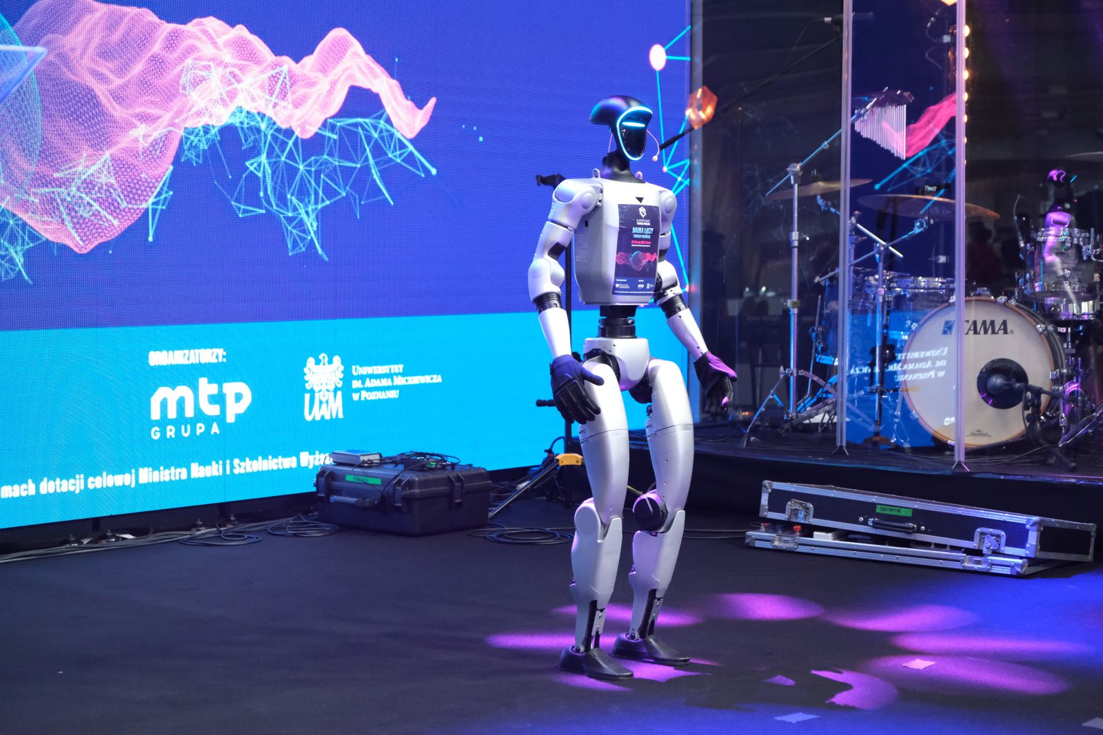 Humanoidalny robot stoi na scenie obok zestawu perkusyjnego, na tle dużego ekranu z kolorową grafiką i logotypami organizatorów Europejskich Targów Nauki.