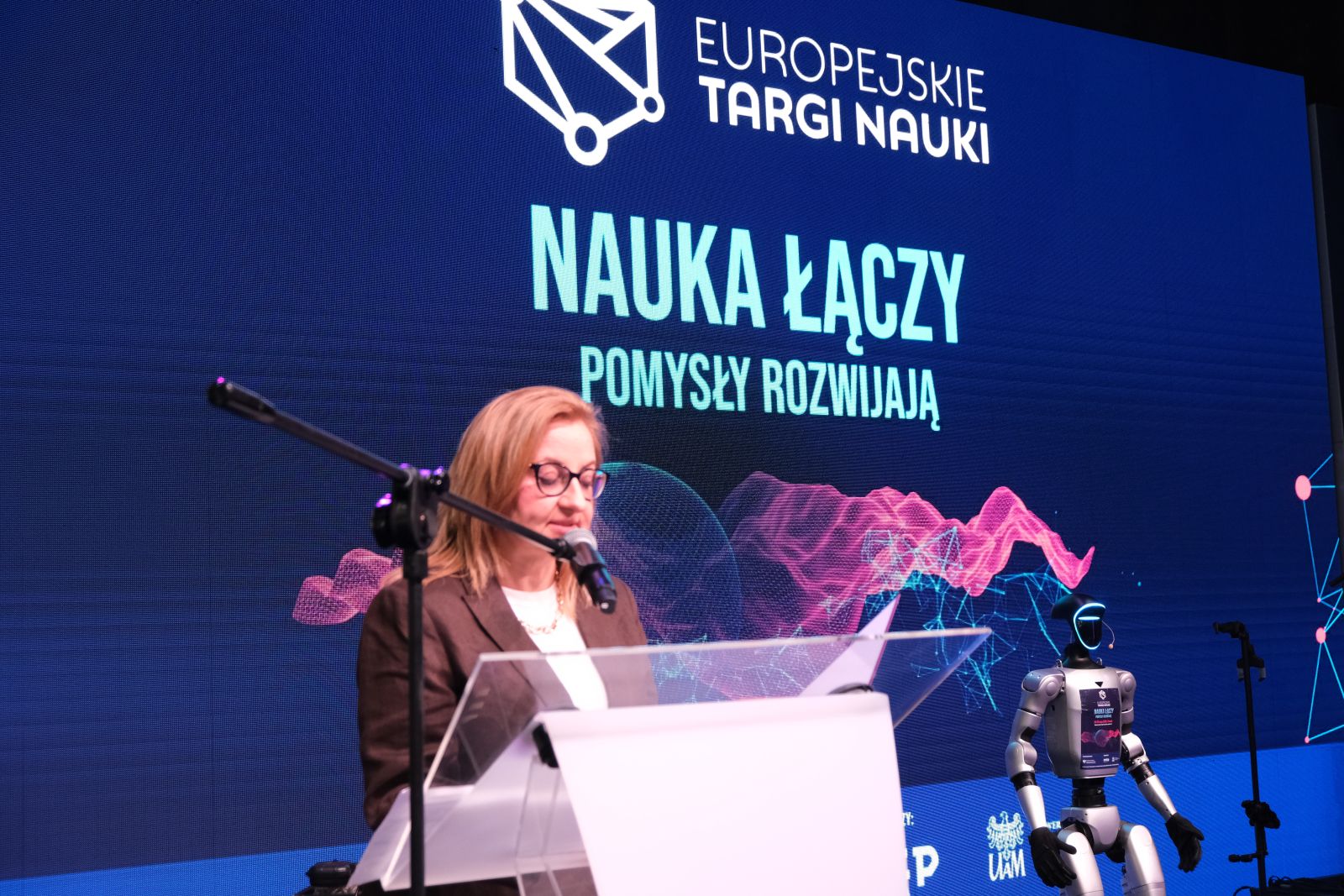 Osoba stoi przy mównicy i odczytuje tekst podczas wydarzenia „Europejskie Targi Nauki”; w tle widoczny jest ekran z hasłem „Nauka łączy, pomysły rozwijają” oraz humanoidalny robot.