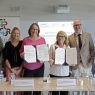 Grupa osób stoi za stołem i prezentuje podpisane dokumenty podczas wydarzenia EPICUR; w tle widoczne są banery oraz ekran z informacją o ceremonii podpisania.