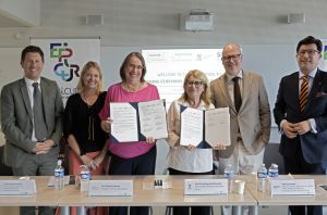 Grupa osób stoi za stołem i prezentuje podpisane dokumenty podczas wydarzenia EPICUR; w tle widoczne są banery oraz ekran z informacją o ceremonii podpisania.