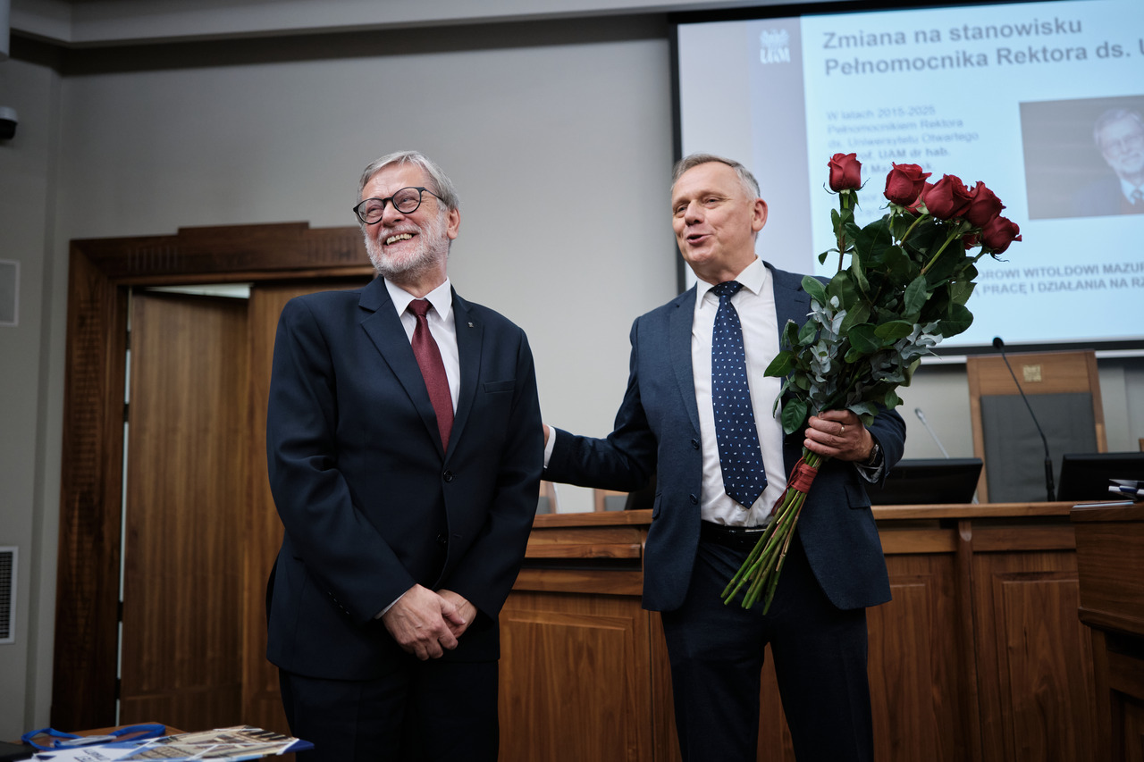Dwaj mężczyźni w garniturach stoją przed salą Uniwersytetu im. Adama Mickiewicza w Poznaniu. Jeden z nich trzyma bukiet czerwonych róż, obaj uśmiechają się.
