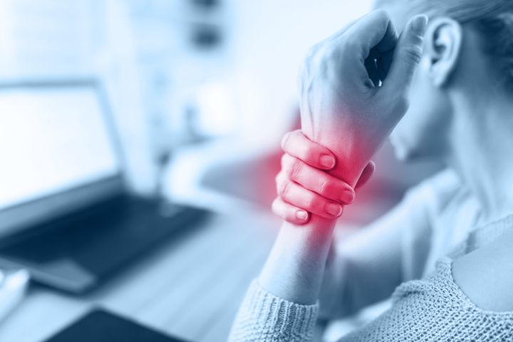 A person sitting at a computer holds their sore wrist with their other hand; the area of pain is marked in red.