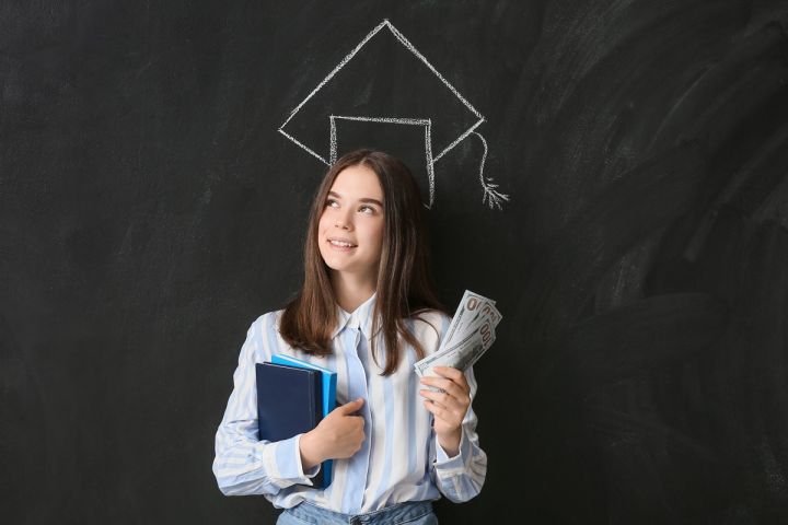 Młoda kobieta trzymająca książki i banknoty, stojąca na tle tablicy z narysowaną czapką absolwencką.