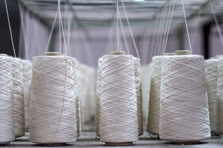 Rzędy białych szpulek z nawiniętą nicią w hali produkcyjnej lub przędzalni, z widocznymi prowadnicami nici u góry.
