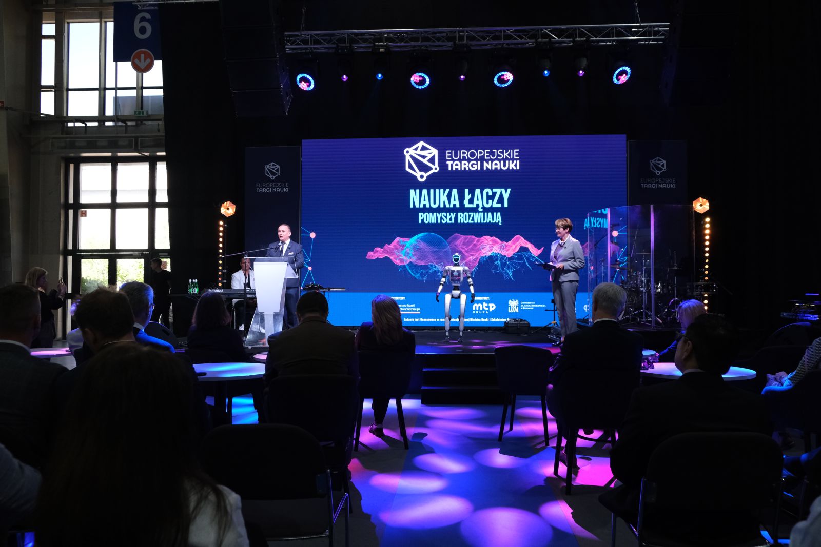 Widok ogólny sceny i publiczności podczas wydarzenia „Europejskie Targi Nauki”; na scenie przemawia osoba przy mównicy, obok stoi robot, a w tle wyświetlone jest hasło „Nauka łączy, pomysły rozwijają”.