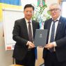 Dwie osoby stoją obok siebie i trzymają zamkniętą teczkę z logotypem UAM podczas oficjalnego spotkania; w tle widoczna jest tablica oraz flaga.