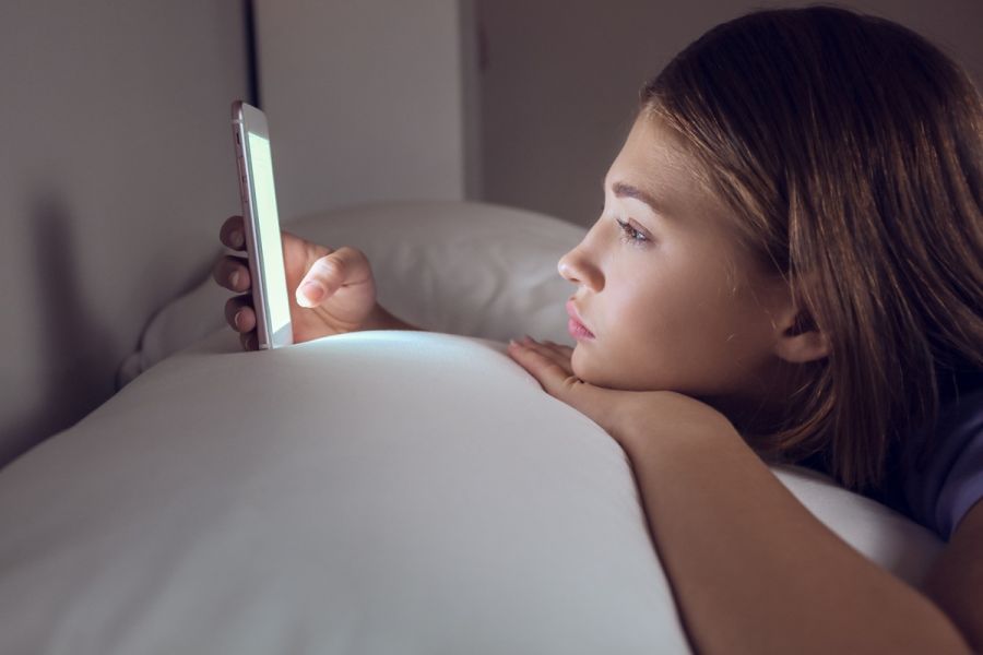 Dziewczyna leży w łóżku po ciemku i patrzy w ekran smartfona, którego światło oświetla jej twarz.