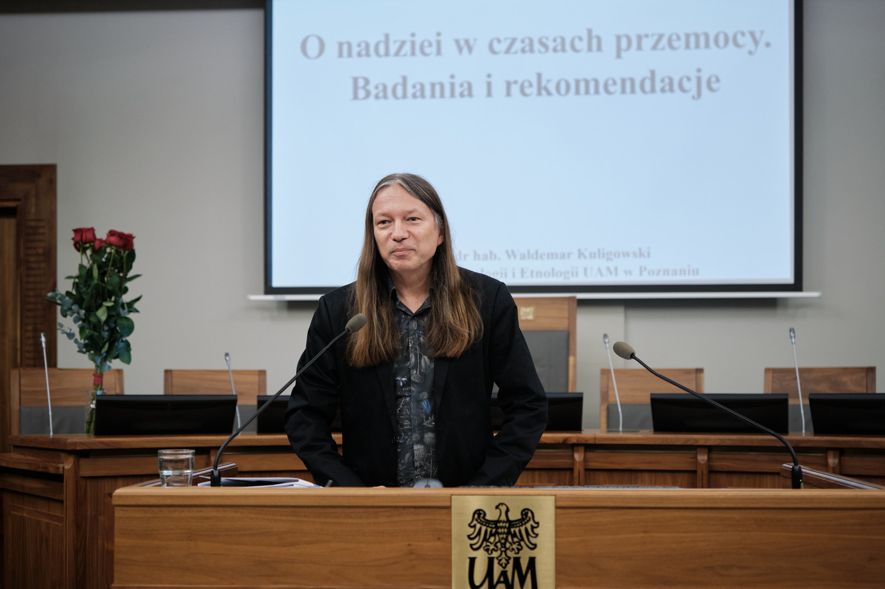 Mężczyzna stoi przy mównicy z logo Uniwersytetu im. Adama Mickiewicza w Poznaniu. W tle ekran z prezentacją