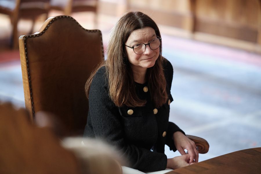 Osoba w okularach siedzi na drewnianym krześle przy stole w eleganckim wnętrzu i uczestniczy w spotkaniu.
